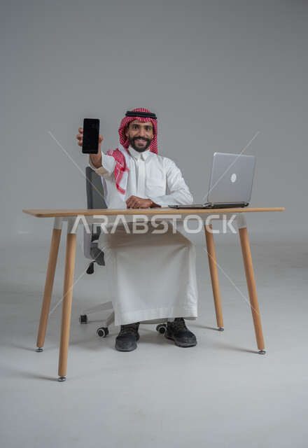 عرض تطبيقات وخدمات الهاتف المحمول, بورتريه لشاب عربي خليجي سعودي يرتدي الثوب التقليدي والشماغ يحمل الجوال بشاشة سوداء فارغة, استخدام الأجهزة والتقنيات الحديثة, التواصل مع الأهل والأصدقاء عبر الأونلاين, خلفية رمادي