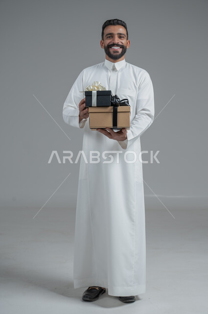 مفهوم الشكر والامتنان، تقديم الهدايا بالاعياد والمناسبات الخاصة، التعبير عن الحب والتقدير والاحترام، بورتريه لرجل عربي سعودي يرتدي الثوب التقليدي يحمل بيديه هدية، الجسم بالطول الكامل، خلفية رمادي