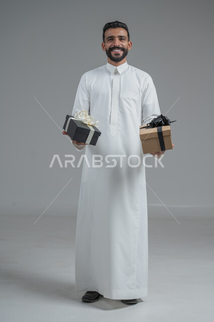 مفهوم الشكر والامتنان، تقديم الهدايا بالاعياد والمناسبات الخاصة، التعبير عن الحب والتقدير والاحترام، بورتريه لرجل عربي سعودي يرتدي الثوب التقليدي يحمل بيديه هدية، الجسم بالطول الكامل، خلفية رمادي