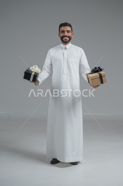 مفهوم الشكر والامتنان، تقديم الهدايا بالاعياد والمناسبات الخاصة، التعبير عن الحب والتقدير والاحترام، بورتريه لرجل عربي سعودي يرتدي الثوب التقليدي يحمل بيديه هدية، الجسم بالطول الكامل، خلفية رمادي