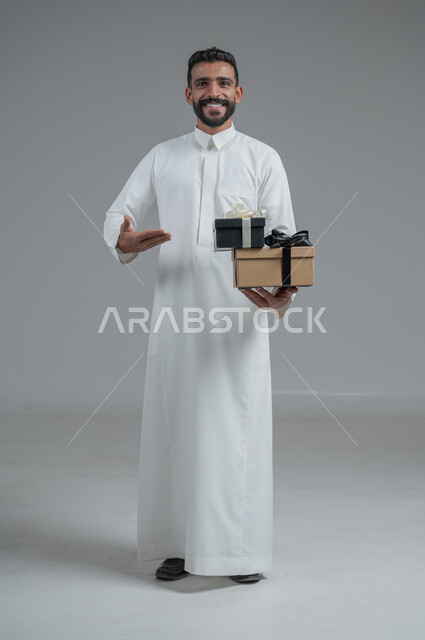 مفهوم الشكر والامتنان، تقديم الهدايا بالاعياد والمناسبات الخاصة، التعبير عن الحب والتقدير والاحترام، بورتريه لرجل عربي سعودي يرتدي الثوب التقليدي يحمل بيديه هدية، الجسم بالطول الكامل، خلفية رمادي