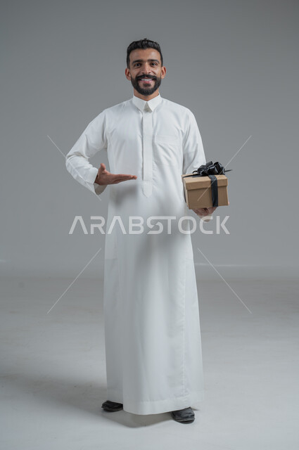 مفهوم الشكر والامتنان، تقديم الهدايا بالاعياد والمناسبات الخاصة، التعبير عن الحب والتقدير والاحترام، بورتريه لرجل عربي سعودي يرتدي الثوب التقليدي يحمل بيديه هدية، الجسم بالطول الكامل، خلفية رمادي