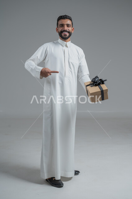 مفهوم الشكر والامتنان، تقديم الهدايا بالاعياد والمناسبات الخاصة، التعبير عن الحب والتقدير والاحترام، بورتريه لرجل عربي سعودي يرتدي الثوب التقليدي يحمل بيديه هدية، الجسم بالطول الكامل، خلفية رمادي