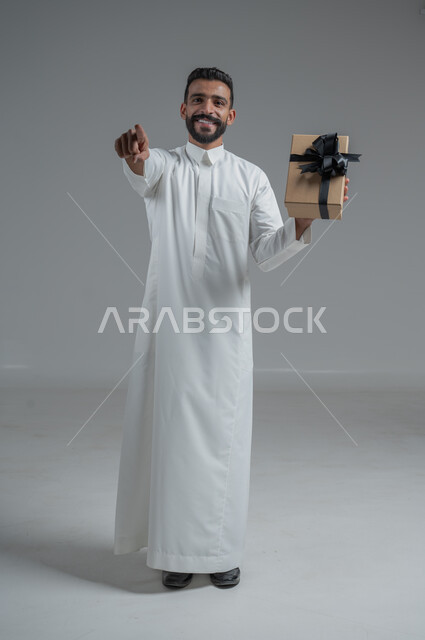The concept of thanks and gratitude, giving gifts on holidays and special occasions, expressing love, appreciation and respect, portrait of a Saudi Arab man wearing a traditional thobe holding a gift in his hands, full-length body, gray background