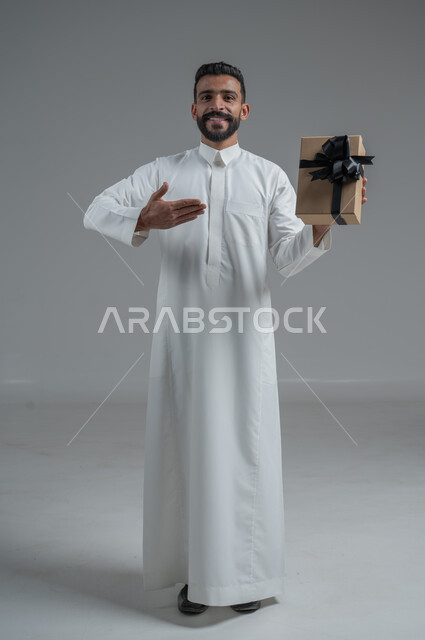 The concept of thanks and gratitude, giving gifts on holidays and special occasions, expressing love, appreciation and respect, portrait of a Saudi Arab man wearing a traditional thobe holding a gift in his hands, full-length body, gray background