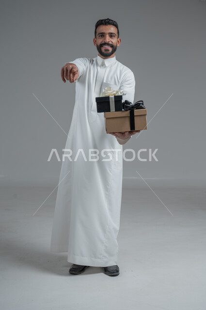 The concept of thanks and gratitude, giving gifts on holidays and special occasions, expressing love, appreciation and respect, portrait of a Saudi Arab man wearing a traditional thobe holding a gift in his hands, full-length body, gray background