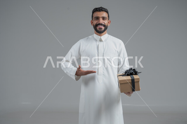 مفهوم الشكر والامتنان، تقديم الهدايا بالاعياد والمناسبات الخاصة، التعبير عن الحب والتقدير والاحترام، بورتريه  مقرب لرجل عربي سعودي يرتدي الثوب التقليدي يحمل بيديه هدية، خلفية رمادي