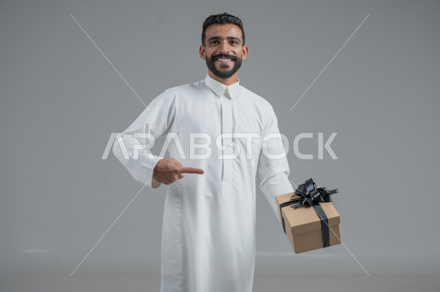 مفهوم الشكر والامتنان، تقديم الهدايا بالاعياد والمناسبات الخاصة، التعبير عن الحب والتقدير والاحترام، بورتريه  مقرب لرجل عربي سعودي يرتدي الثوب التقليدي يحمل بيديه هدية، خلفية رمادي