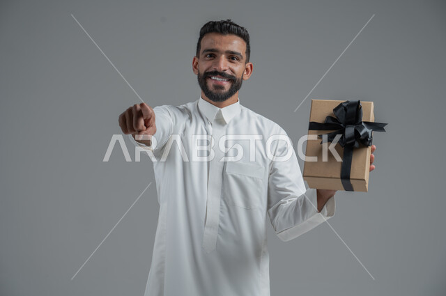 مفهوم الشكر والامتنان، تقديم الهدايا بالاعياد والمناسبات الخاصة، التعبير عن الحب والتقدير والاحترام، بورتريه  مقرب لرجل عربي سعودي يرتدي الثوب التقليدي يحمل بيديه هدية، خلفية رمادي