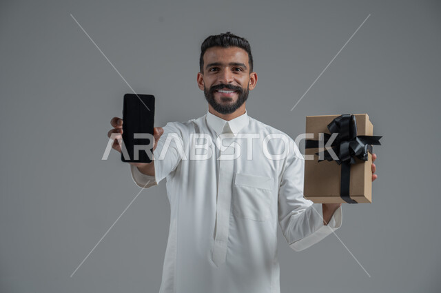 مفهوم الشكر والامتنان، تقديم الهدايا بالاعياد والمناسبات الخاصة، التعبير عن الحب والتقدير والاحترام، بورتريه  مقرب لرجل عربي سعودي يرتدي الثوب التقليدي يحمل بيديه هدية وهاتف محمول بخلفية سوادء، خلفية رمادي
