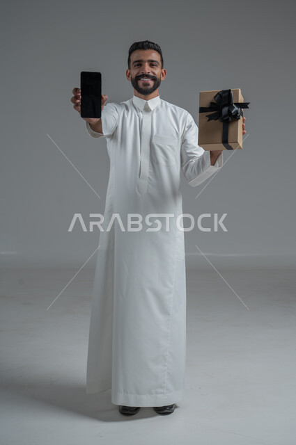 مفهوم الشكر والامتنان، تقديم الهدايا بالاعياد والمناسبات الخاصة، التعبير عن الحب والتقدير والاحترام، بورتريه  لرجل عربي سعودي يرتدي الثوب التقليدي يحمل بيديه هدية وهاتف محمول بخلفية سوادء، صورة الجسم بالطول الكامل, خلفية رمادي