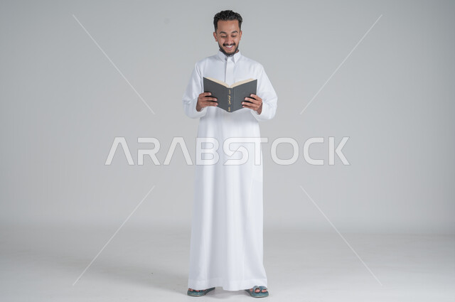  مفهوم التمعن والتركيز، قضاء الوقت بالقراءة والتعلم والثقافة، بورتريه لرجل عربي سعودي خليجي يرتدي الثوب التقليدي يحمل بيديه كتاب، الجسم بالطول الكامل، خلفية رمادي