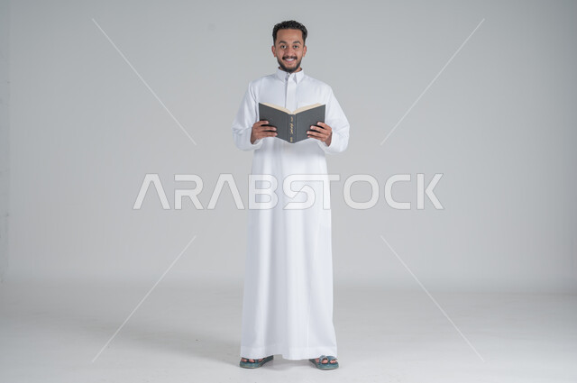 The concept of contemplation and concentration, spending time reading, learning and culture, portrait of a Saudi Arabian Gulf man wearing traditional thobe holding a book in his hands, full body, gray background