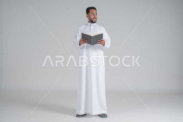  مفهوم التمعن والتركيز، قضاء الوقت بالقراءة والتعلم والثقافة، بورتريه لرجل عربي سعودي خليجي يرتدي الثوب التقليدي يحمل بيديه كتاب، الجسم بالطول الكامل، خلفية رمادي