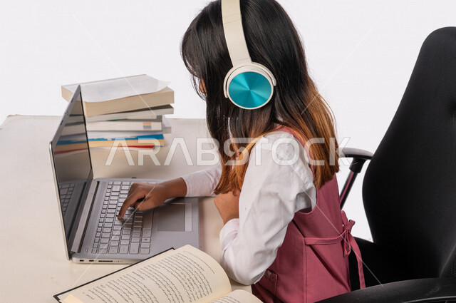 A Saudi Arabian Gulf schoolgirl wearing a school uniform and a headset, sitting in front of a laptop, e-learning, distance study, following up on lessons and assignments online, new semester, education in the Kingdom of Saudi Arabia