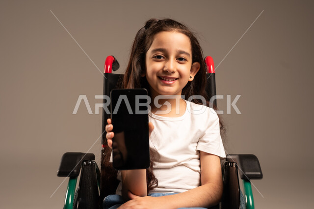 عرض تطبيقات لأصحاب الاحتياجات الخاصة عبر الهاتف المحمول، بورتريه مقرب لفتاة عربية خليجية سعودية مبتسمة تجلس على كرسي متحرك تحمل بيدها جوال بشاشة سوداء فارغة، تسهيل حياة ذوي الهمم باستخدام التكنولوجيا، تعليم المعاقين على الأجهزة التقنية, خلفية رمادية