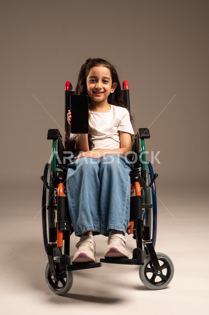 عرض تطبيقات لأصحاب الاحتياجات الخاصة عبر الهاتف المحمول، بورتريه لفتاة عربية خليجية سعودية مبتسمة تجلس على كرسي متحرك تحمل بيدها جوال بشاشة سوداء فارغة، تسهيل حياة ذوي الهمم باستخدام التكنولوجيا، تعليم المعاقين على الأجهزة التقنية, خلفية رمادية