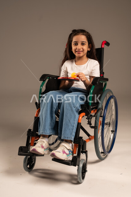 النظر الى الكاميرا بايماءات وتعابير السعادة والسرور, تنمية ودعم المجتمع السعودي للمواهب الرياضية، التمكين الاجتماعي للمعاقين, بورتريه لفتاة مقعدة عربية خليجية سعودية مبتسمة تجلس على كرسي متحرك تحمل بيدها مضرب وكرة الطاولة، خلفية رمادية