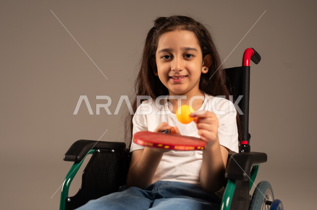 النظر الى الكاميرا بايماءات وتعابير السعادة والسرور, تنمية ودعم المجتمع السعودي للمواهب الرياضية، التمكين الاجتماعي للمعاقين, بورتريه لفتاة مقعدة عربية خليجية سعودية مبتسمة تجلس على كرسي متحرك تحمل بيدها مضرب وكرة الطاولة، خلفية رمادية