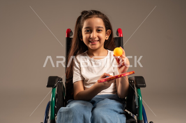 النظر الى الكاميرا بايماءات وتعابير السعادة والسرور, تنمية ودعم المجتمع السعودي للمواهب الرياضية، التمكين الاجتماعي للمعاقين, بورتريه لفتاة مقعدة عربية خليجية سعودية مبتسمة تجلس على كرسي متحرك تحمل بيدها مضرب وكرة الطاولة، خلفية رمادية