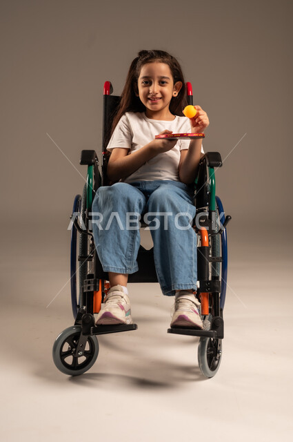 النظر الى الكاميرا بايماءات وتعابير السعادة والسرور, تنمية ودعم المجتمع السعودي للمواهب الرياضية، التمكين الاجتماعي للمعاقين, بورتريه لفتاة مقعدة عربية خليجية سعودية مبتسمة تجلس على كرسي متحرك تحمل بيدها مضرب وكرة الطاولة، خلفية رمادية