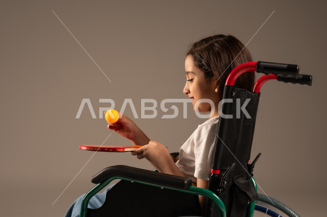 تنمية ودعم المجتمع السعودي للمواهب الرياضية، التمكين الاجتماعي للمعاقين, بورتريه من الجانب لفتاة مقعدة عربية خليجية سعودية مبتسمة تجلس على كرسي متحرك تحمل بيدها مضرب وكرة الطاولة بايماءات وتعابير السعادة والسرور, خلفية رمادية