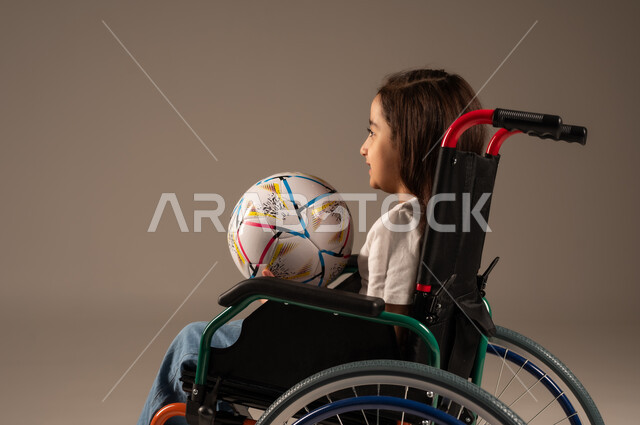  دعم وتنمية المجتمع السعودي للمواهب الرياضية، التمكين الاجتماعي للمعاقين, بورتريه من الجانب لفتاة مقعدة عربية خليجية سعودية مبتسمة تجلس على كرسي متحرك تحمل بيدها كرة تنظر الى شيء ما بايماءات السعادة والبشاشة بفوز الفريق المفضل, خلفية رمادية