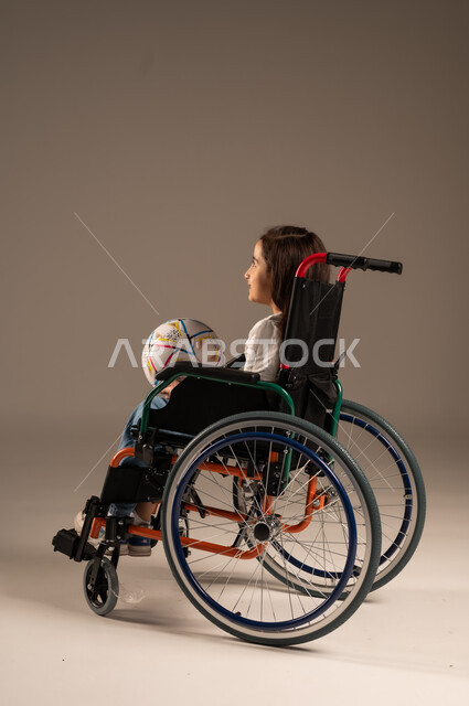 Support and development of Saudi society for sports talents, social empowerment of the disabled, side portrait of a smiling Saudi Arabian Gulf girl sitting in a wheelchair holding a ball in her hand looking at the camera with gestures of happiness and cheerfulness for the victory of her favorite team, gray background