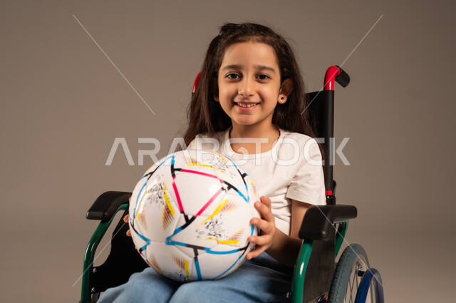 Support and development of Saudi society for sports talents, social empowerment of the disabled, close-up portrait of a smiling Saudi Arabian Gulf disabled girl sitting in a wheelchair holding a ball in her hand looking at the camera with gestures of happiness and cheerfulness for the victory of her favorite team, gray background
