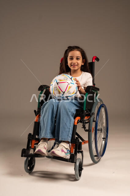 Support and development of Saudi society for sports talents, social empowerment of the disabled, portrait of a smiling Saudi Arabian Gulf disabled girl sitting in a wheelchair holding a ball in her hand looking at the camera with gestures of happiness and cheerfulness for the victory of her favorite team, gray background