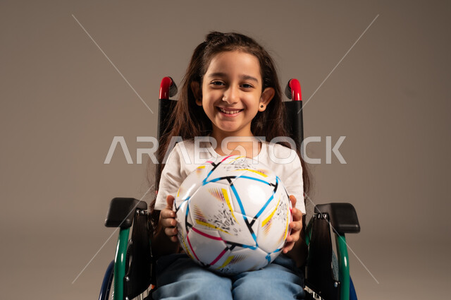 Support and development of Saudi society for sports talents, social empowerment of the disabled, close-up portrait of a smiling Saudi Arabian Gulf disabled girl sitting in a wheelchair holding a ball in her hand looking at the camera with gestures of happiness and cheerfulness for the victory of her favorite team, gray background