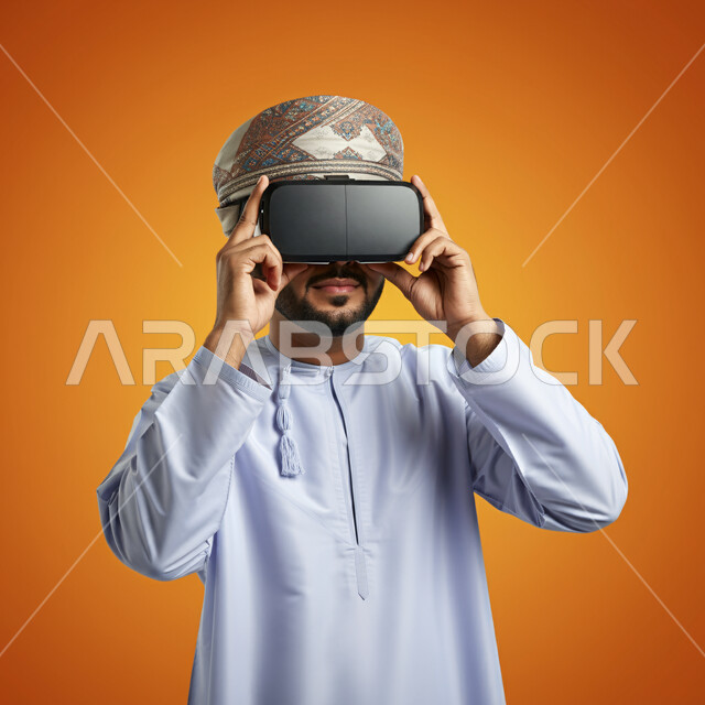 Integration and simulation of the imaginary world, technical and technological development, fun and entertainment by watching through virtual reality glasses, close-up portrait of a young Gulf Arab Omani man wearing a dishdasha and turban looking through 3D glasses and holding them in his hand, orange background