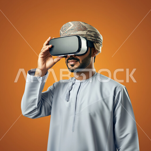  الاندماج ومحاكاة العالم الخيالي ،التطور التقني والتكنولوجي, المتعة و التسلية بالمشاهدة عبر نظارة الواقع الافتراضي، بورتريه مقرب لشاب عربي خليجي عماني يرتدي الدشداشه والعمامة ينظر من خلال نظارات ثلاثية الأبعاد ويمسكها بيده, خلفيه برتقالية