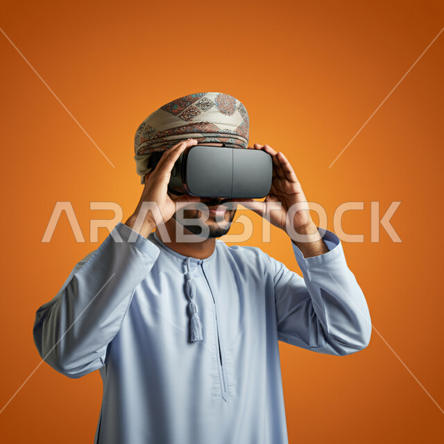 Technical and technological development, fun and entertainment by watching through virtual reality glasses, close-up portrait of a young Gulf Arab Omani man wearing a dishdasha and turban looking through 3D glasses and holding them in his hand, integration and simulation of the imaginary world, orange background