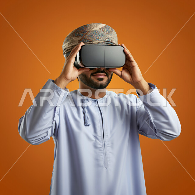 التطور التقني والتكنولوجي, المتعة و التسلية بالمشاهدة عبر نظارة الواقع الافتراضي، بورتريه مقرب لشاب عربي خليجي عماني يرتدي الدشداشه والعمامة ينظر من خلال نظارات ثلاثية الأبعاد ويمسكها بيده, الاندماج ومحاكاة العالم الخيالي ، خلفيه برتقالية