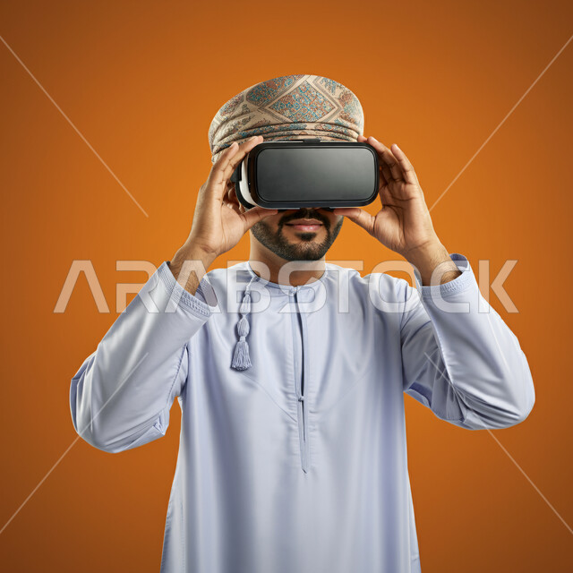 Technical and technological development, fun and entertainment by watching through virtual reality glasses, close-up portrait of a young Gulf Arab Omani man wearing a dishdasha and turban looking through 3D glasses and holding them in his hand, integration and simulation of the imaginary world, orange background