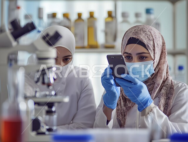 استخدام أجهزة الفحص المتطورة لتحليل العينات المختلفة، مختبرات الفحوصات الطبية والتحاليل الكيمائية، معمل الصناعات الدوائية، امرأتان عربيتان خليجيتان سعوديتان محجبتان ترتديان المعطف والكمامة تقوم بالبحث حول العينة والنظر في المجهر، إجراء التجارب والبحوث العلمية الحيوية