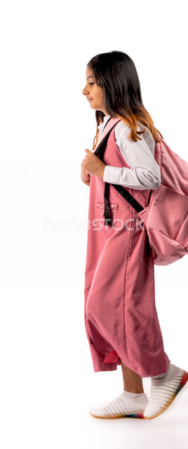 Photograph from the side of a Saudi Arabian Gulf schoolgirl wearing a school uniform and school bag, ready to go to school, preparing for the new semester, education in Saudi Arabia, white background