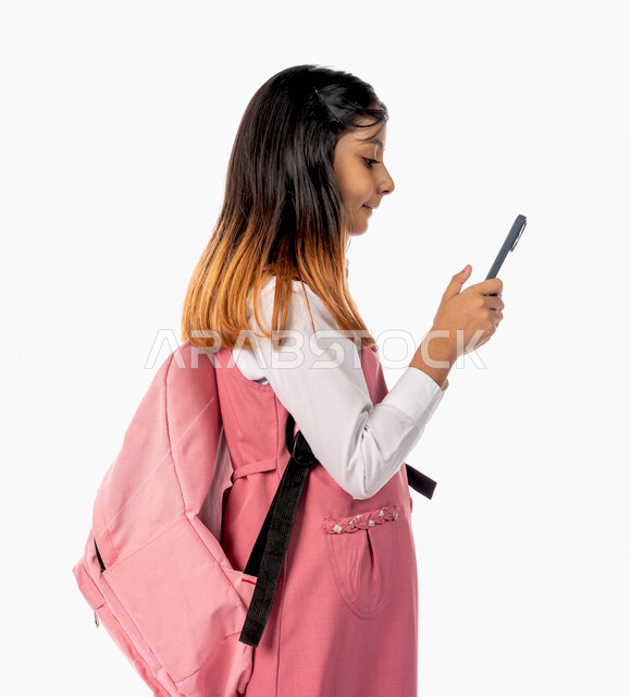 A Saudi Arabian Gulf schoolgirl wearing a school uniform, using a mobile phone, e-learning, remote study, following up on lessons and assignments online, new semester, education in Saudi Arabia, white background