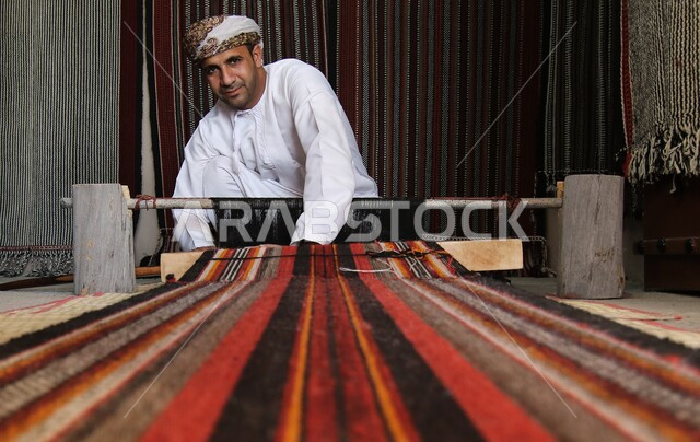 صناعات وحرف يدوية شعبية تقليدية، المهن العمانية القديمة في الغزل وصناعة السجاد، رجل يرتدي العمامة والدشداشه يقوم بحياكة الأنسجة والقماش، النظر إلى الكاميرا بإيماءات الفخر والسرور، استخدام الخيوط الملونة، الفنون والمهارات التراثية،