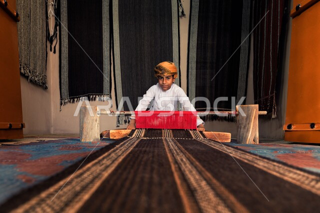 Traditional folk crafts and industries, ancient Omani professions in spinning and carpet making, a boy wearing a turban and dishdasha weaving fabrics and cloth, looking at the camera with gestures of pride and pleasure, using colored threads, traditional arts and skills,