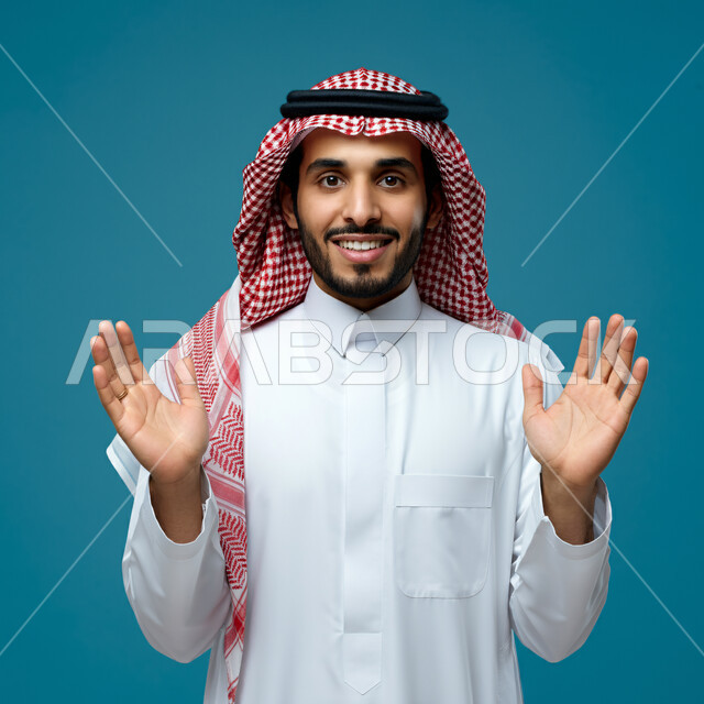 تحديد الحجم والكتله، بورتريه مقرب لرجل عربي خليجي سعودي مبتسم يرتدي الثوب التقليدي والشماغ يقف باستقامة يقوم برفع كفيه للأعلى لتحجيم شيء ما، النظر الى الكاميرا بتعابير الفرح والسرور، مفهوم تقديم وعرض عنصر معين، خلفية زرقاء