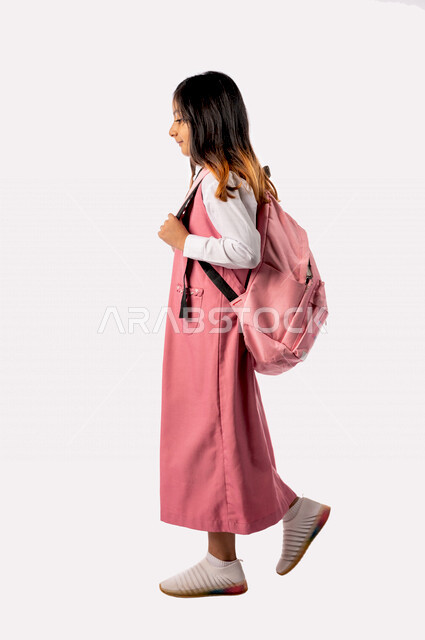 تصوير من الجانب لتلميذة عربية خليجية سعودية ترتدي الزي المدرسي والحقيبة المدرسية  ،مستعدة للذهاب الى المدرسة ، الاستعداد للفصل الدراسي الجديد، التعليم في المملكة العربية السعودية ، خلفية بيضاء