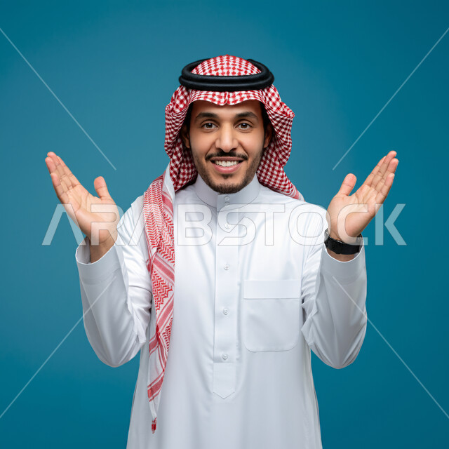 تحديد الحجم والكتله، بورتريه مقرب لرجل عربي خليجي سعودي مبتسم يرتدي الثوب التقليدي والشماغ يقف باستقامة يقوم برفع كفيه للأعلى لتحجيم شيء ما، النظر الى الكاميرا بتعابير الفرح والسرور، مفهوم تقديم وعرض عنصر معين، خلفية زرقاء