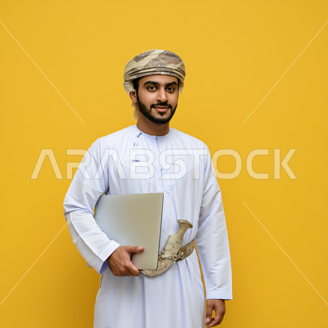  مفهوم تسهيل الاعمال والقدرة على إنجاز المهام عن بعد، استخدام التقنيات الحديثة والأجهزة المتطورة ، بورتريه لشاب عربي خليجي عماني يرتدي الدشداشة والعمامة يمسك بيده حاسوب محمول، النظر الى الكاميرا بتعابير الفخر والسرور، خلفية صفراء