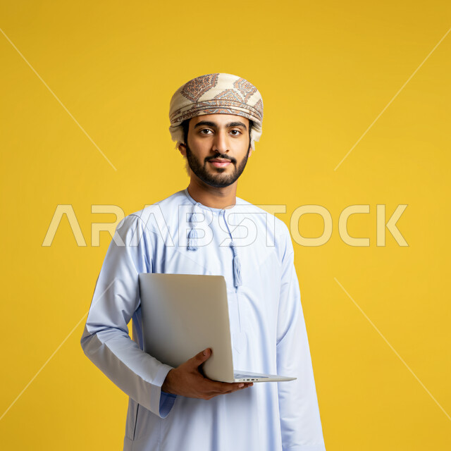  مفهوم تسهيل الاعمال والقدرة على إنجاز المهام عن بعد، استخدام التقنيات الحديثة والأجهزة المتطورة ، بورتريه لشاب عربي خليجي عماني يرتدي الدشداشة والعمامة يمسك بيده حاسوب محمول، النظر الى الكاميرا بتعابير الفخر والسرور، خلفية صفراء