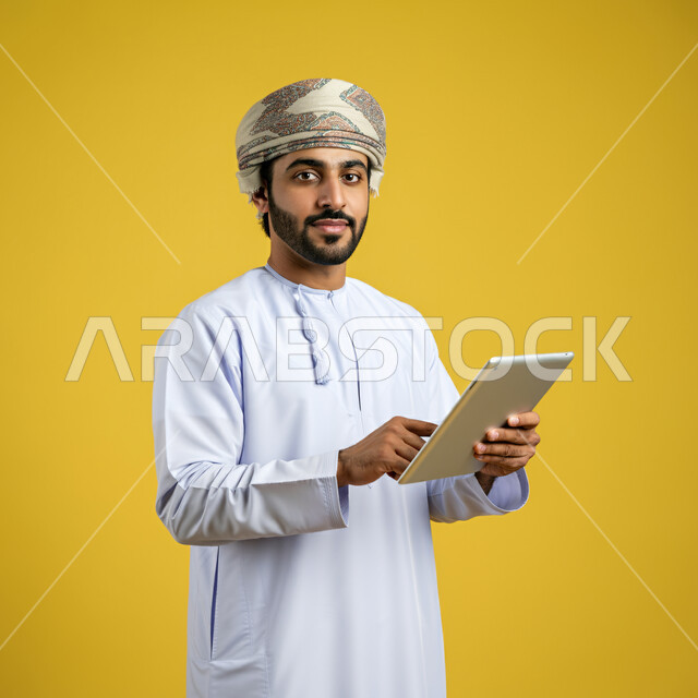 استخدام التكنولوجيا والتقنيات الحديثة, تصفح مواقع التواصل الاجتماعي، متابعة الأفلام والمسلسلات الممتعة، المحادثة مع الأهل والأصدقاء، بورتريه مقرب لرجل عربي خليجي عماني يرتدي الدشداشة والعمامة يحمل بيده جهاز التابلت، النظر الى الكاميرا بتعابير الفرح والسرور، خلفية صفراء