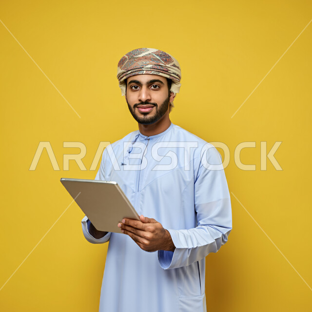 استخدام التكنولوجيا والتقنيات الحديثة, تصفح مواقع التواصل الاجتماعي، متابعة الأفلام والمسلسلات الممتعة، المحادثة مع الأهل والأصدقاء، بورتريه مقرب لرجل عربي خليجي عماني يرتدي الدشداشة والعمامة يحمل بيده جهاز التابلت، النظر الى الكاميرا بتعابير الفرح والسرور، خلفية صفراء