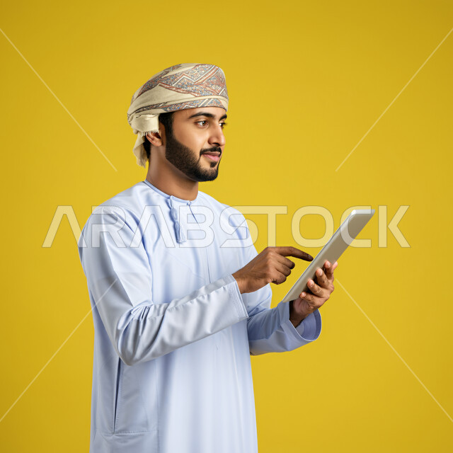 استخدام التكنولوجيا والتقنيات الحديثة, تصفح مواقع التواصل الاجتماعي، متابعة الأفلام والمسلسلات الممتعة، المحادثة مع الأهل والأصدقاء، بورتريه مقرب لرجل عربي خليجي عماني يرتدي الدشداشة والعمامة يحمل بيده جهاز التابلت، النظر بعيدا الى مكان ما بتعابير السرور، خلفية صفراء