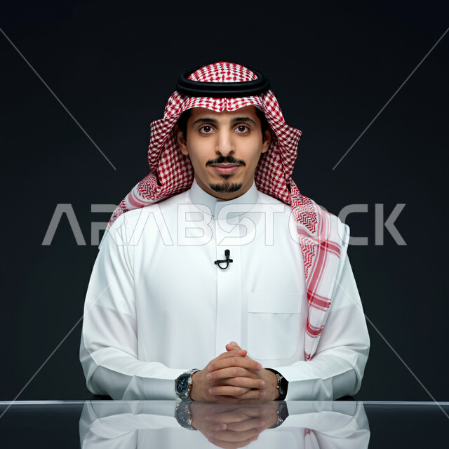 تقديم الأخبار السعودية والعالمية ومستجداتها، وسائل الإعلام التلفزيونية، دمج البيئة العملية مع التقنية والتكنولوجيا، بورتريه مقرب لمذيع عربي سعودي يرتدي الثوب التقليدي والشماغ ينظر إلى الكاميرا بإيماءات الجدية، استخدام تقنية الجرافيكس في تقديم اخبارمحلية و اقليمية، خلفية سوداء